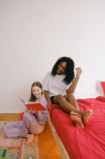 Full body of cheerful barefoot diverse female friends reading interesting book together in light bedroom with red blanket on bed at home